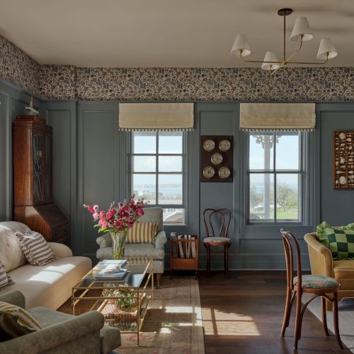 A cozy living room with vintage decor, blue walls, large windows, and assorted furniture including sofas and chairs.