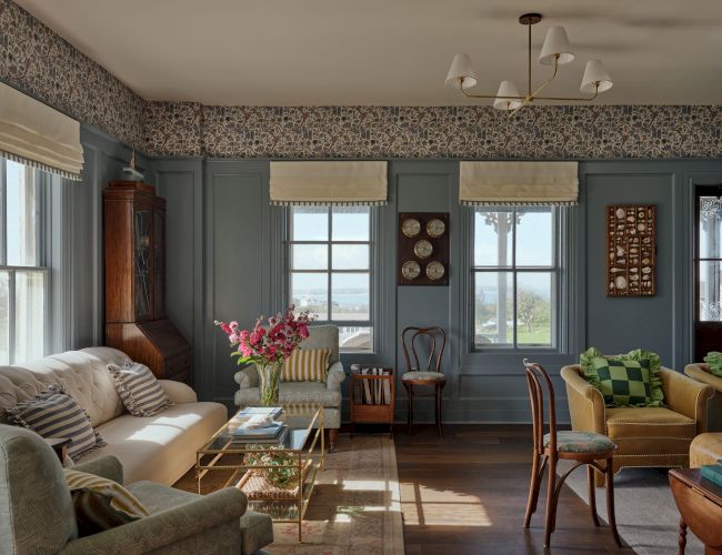 A cozy living room with vintage decor, blue walls, large windows, and assorted furniture including sofas and chairs.