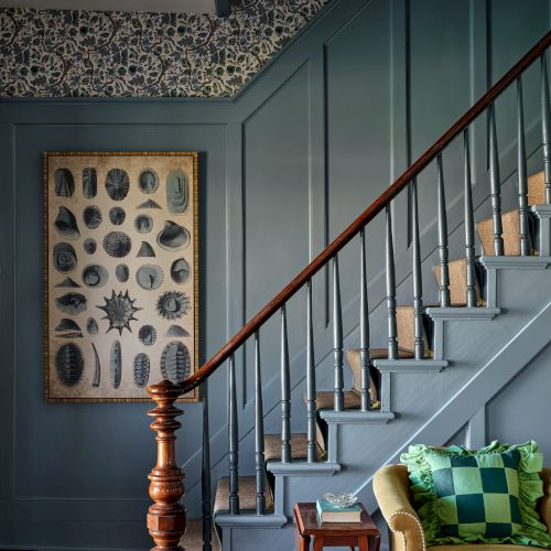 A staircase with blue paneling, a botanical artwork, a wooden railing, and a yellow armchair with a green cushion beside a small table.