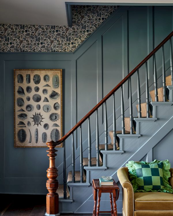 A staircase with blue paneling, a botanical artwork, a wooden railing, and a yellow armchair with a green cushion beside a small table.