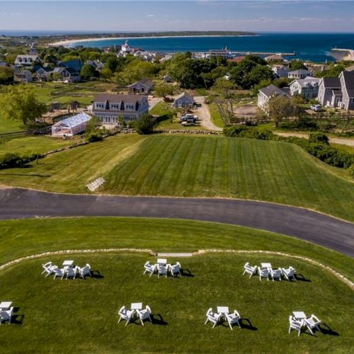 A scenic coastal town with neatly arranged white outdoor chairs on a green lawn, charming houses, and a view of the sea and headland in the background.