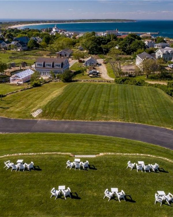 A scenic coastal town with neatly arranged white outdoor chairs on a green lawn, charming houses, and a view of the sea and headland in the background.