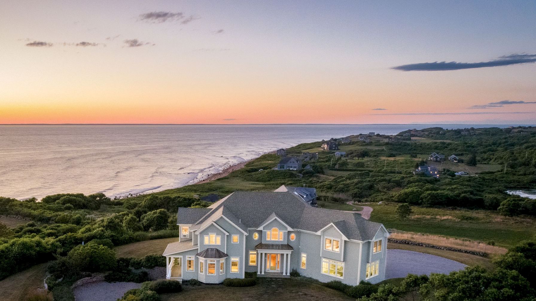 A large coastal house at sunset, with a winding driveway and surrounding greenery, overlooking the ocean along a scenic shoreline.