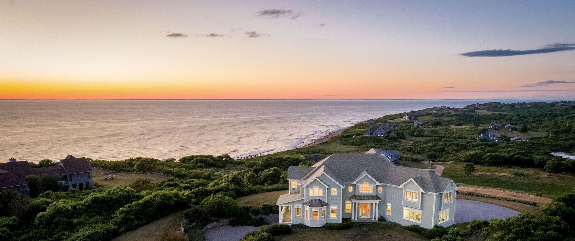 A large coastal house at sunset, with a winding driveway and surrounding greenery, overlooking the ocean along a scenic shoreline.