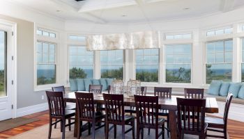 A spacious dining room with a long table, several chairs, large windows, and a modern ceiling light.