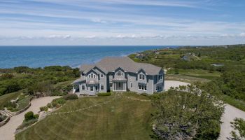 A large blue house overlooks the ocean, surrounded by a manicured lawn, trees, and a driveway, under a blue sky with wispy clouds.