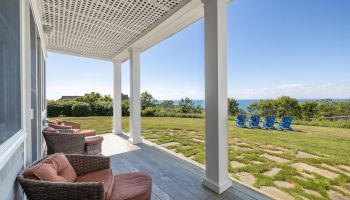 A porch with wicker chairs overlooks a grassy yard and blue Adirondack chairs, with the ocean in the background under a clear sky.