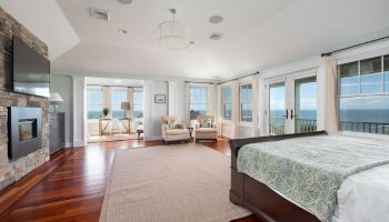 The image shows a spacious bedroom with hardwood floors, a fireplace, large windows, and a seating area with a view.