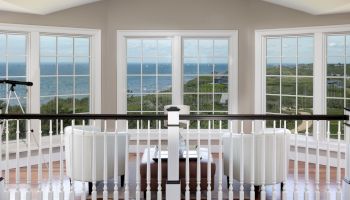 A room with large windows, white chairs, and a table overlooks a scenic ocean view, with a telescope in the corner.