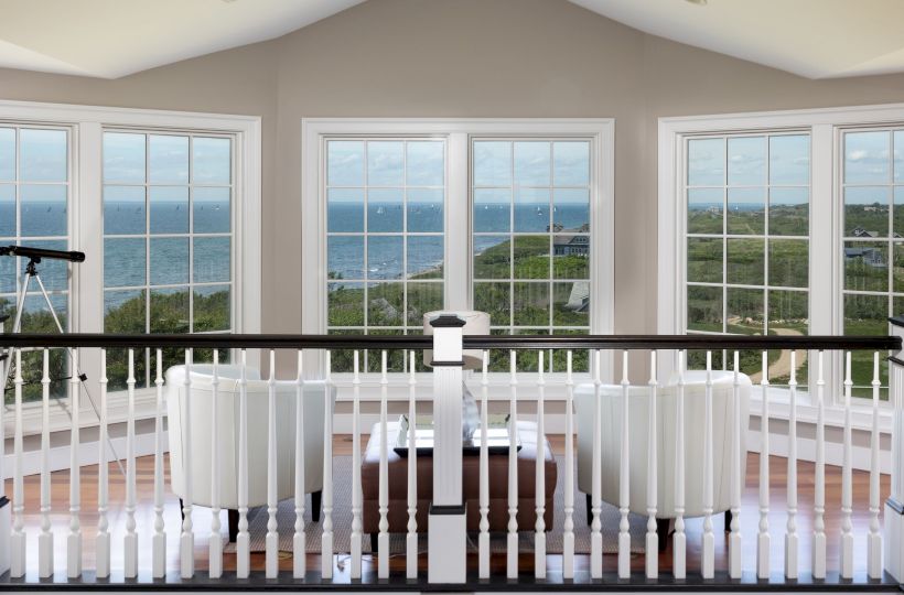 A room with large windows, white chairs, and a table overlooks a scenic ocean view, with a telescope in the corner.