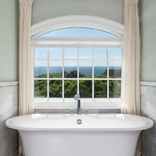 A freestanding bathtub is in front of a large window, offering a scenic view of greenery and sky, with light curtains on either side.