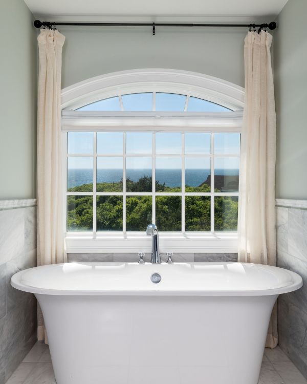 A freestanding bathtub is in front of a large window, offering a scenic view of greenery and sky, with light curtains on either side.