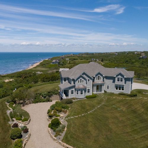 A large house sits by the ocean with a curved driveway, surrounded by greenery and a vast lawn under a blue sky.