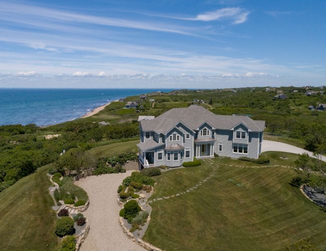 A large house sits by the ocean with a curved driveway, surrounded by greenery and a vast lawn under a blue sky.