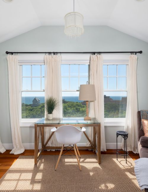 A bright room with large windows, a desk, a chair, and a lounge chair. The view outside is of greenery and a clear sky.