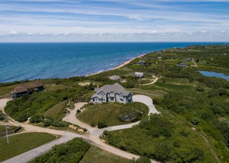 Aerial view of a coastal landscape with a large house, winding roads, greenery, and the ocean in the background under a blue sky.