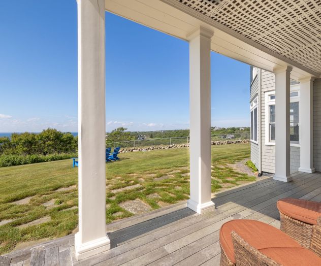 A coastal porch with wicker furniture overlooks a grassy lawn and ocean view, framed by columns under a blue sky with distant trees.