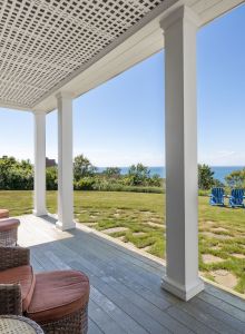 A porch with wicker chairs overlooks a grassy yard with blue chairs, offering a view of the sea on a clear, sunny day.
