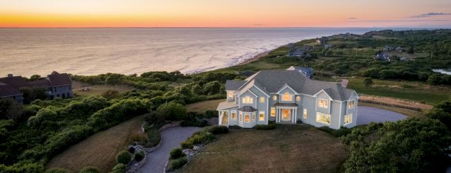 A large coastal house on a spacious plot overlooks a scenic ocean sunset, with a winding driveway and lush greenery surrounding it.