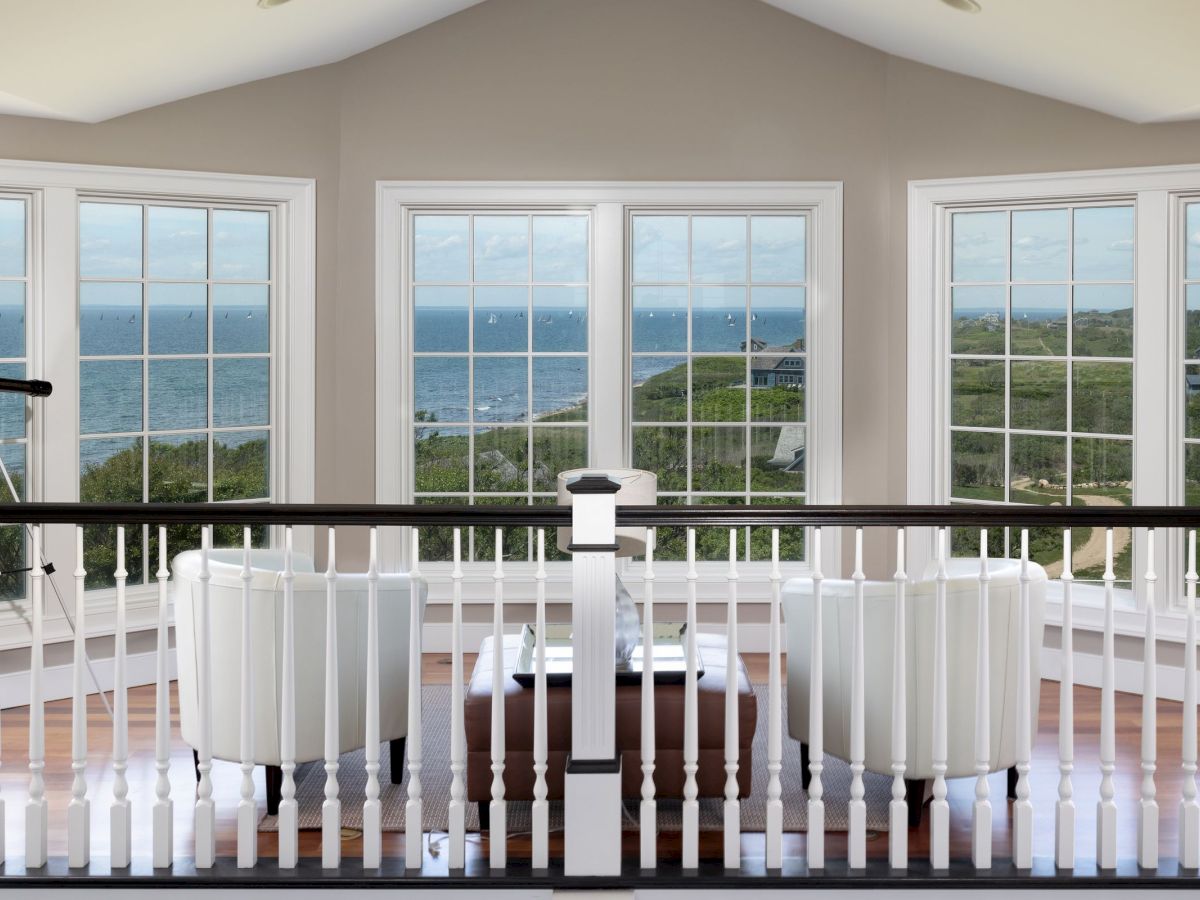 A room with large windows offers an ocean view, featuring two white chairs, a small table, and a telescope on the left.