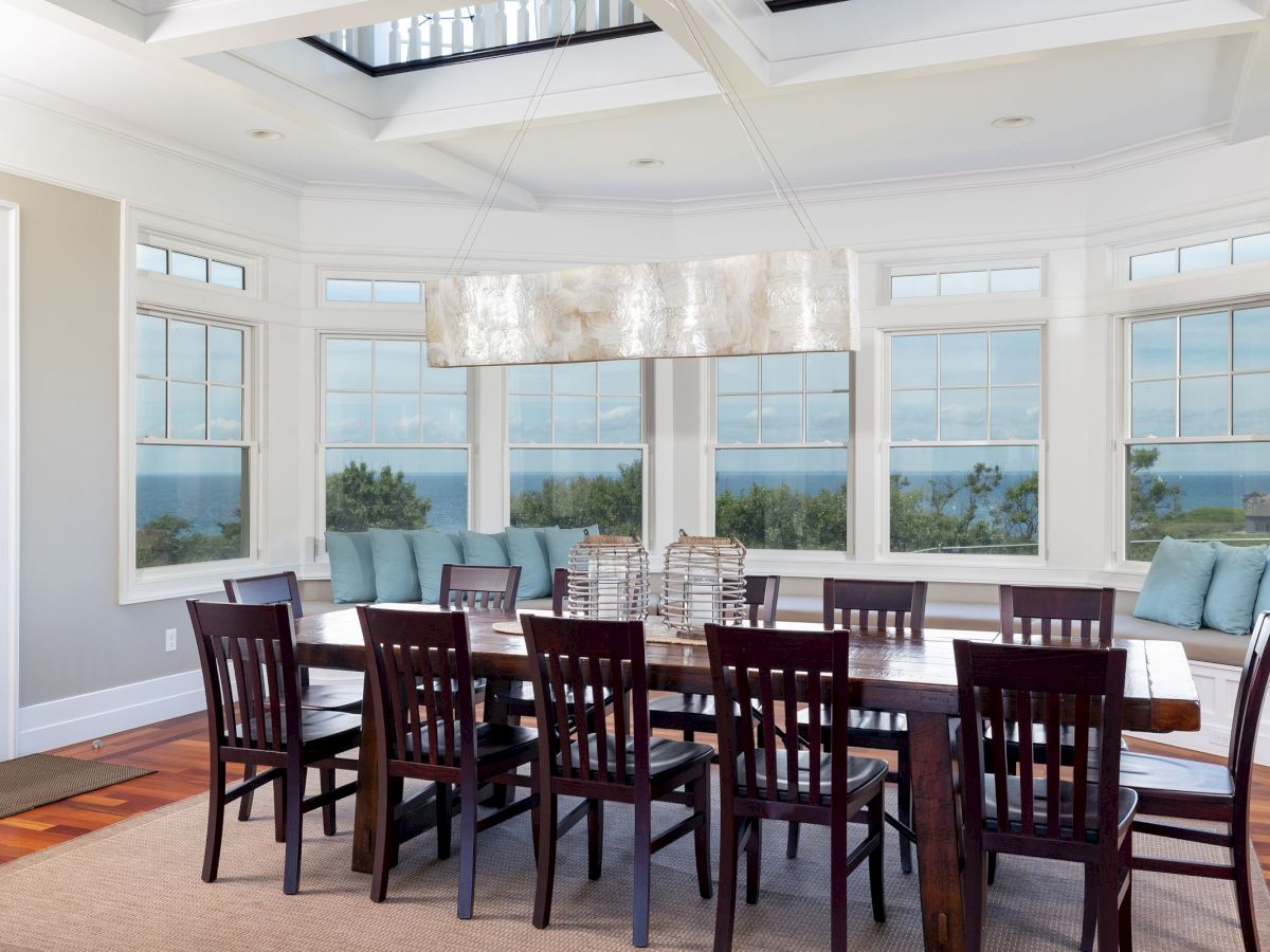 A dining room with a large wooden table, chairs, a modern light fixture, and large windows offering an ocean view.