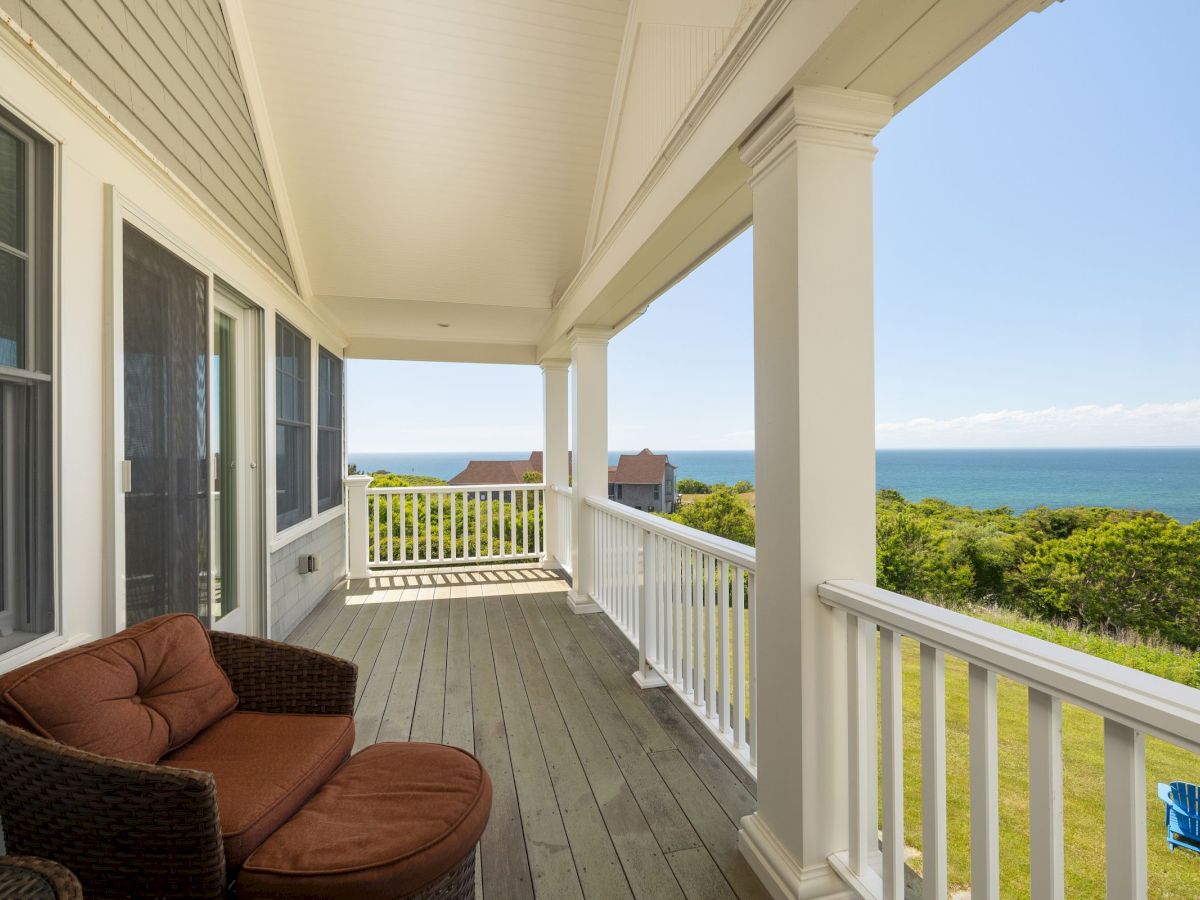 A porch with a wicker chair overlooks a scenic view of the ocean and greenery, under a clear blue sky, creating a serene atmosphere.