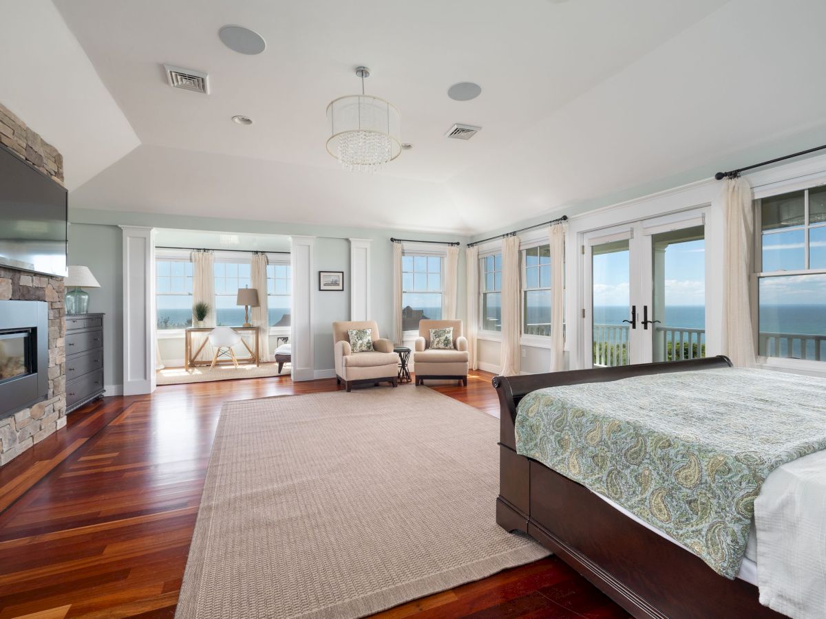 A spacious bedroom with a large bed, seating area, fireplace, TV, and ocean views from glass doors and windows.