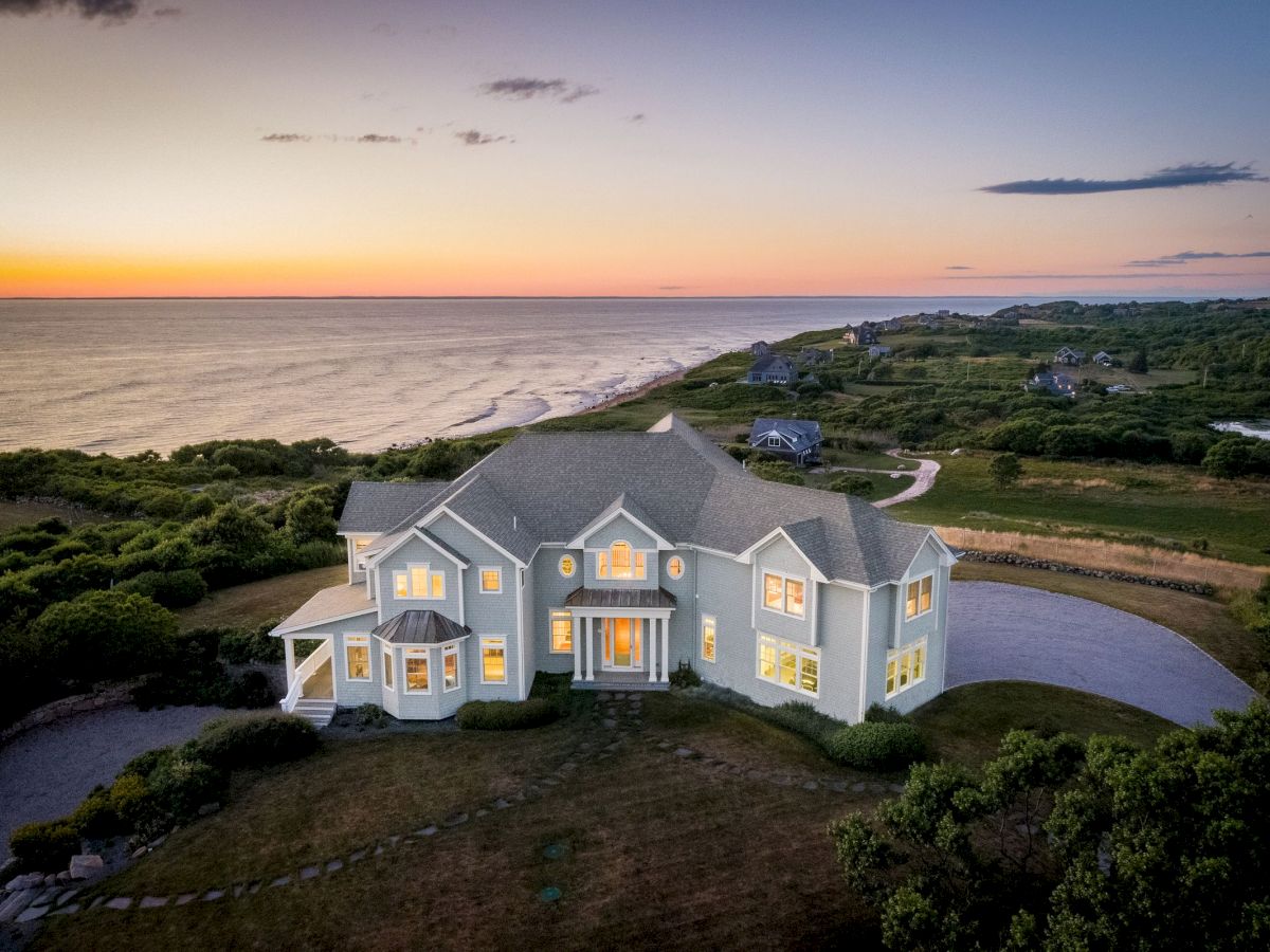 A coastal home at sunset with glowing windows, surrounded by greenery, overlooking the ocean and a winding road.