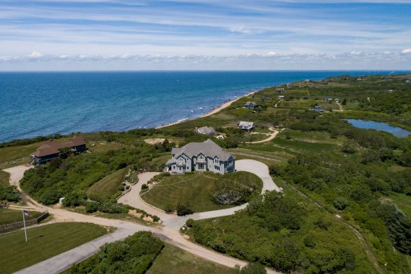 A coastal landscape with a large house, winding roads, green vegetation, and the ocean in the background under a blue sky.