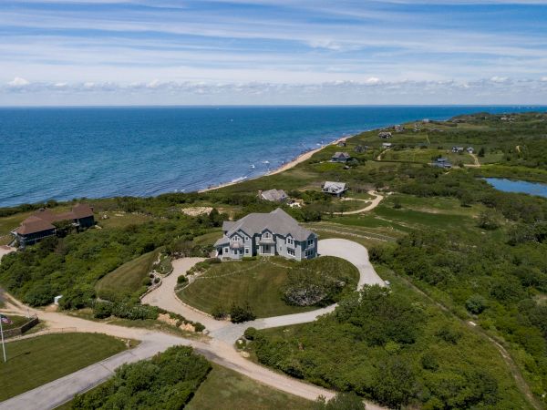 A coastal landscape with a large house, winding roads, green vegetation, and the ocean in the background under a blue sky.