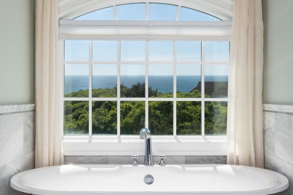 A white bathtub is set against a window with white curtains, offering a view of greenery and the sky outside.