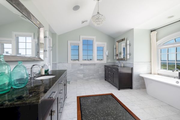 This image shows a bright bathroom with a bathtub, large mirrors, two sinks, and windows. There's a chandelier overhead.