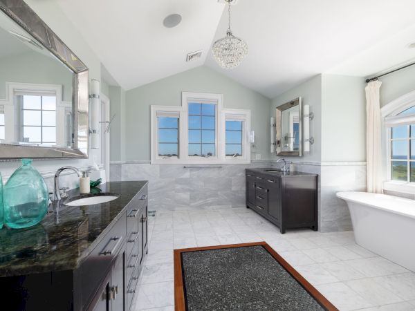 This image shows a bright bathroom with a bathtub, large mirrors, two sinks, and windows. There's a chandelier overhead.