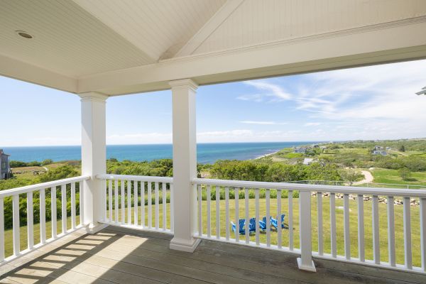 A porch with white railings offers a view of the ocean, grassy fields, and a winding path under a partly cloudy sky.