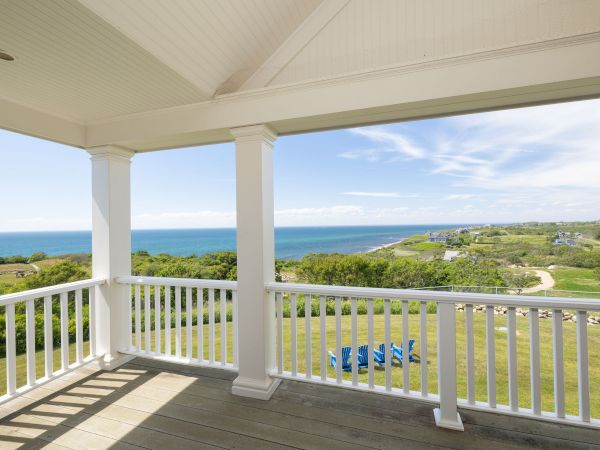A porch with white railings offers a view of the ocean, grassy fields, and a winding path under a partly cloudy sky.