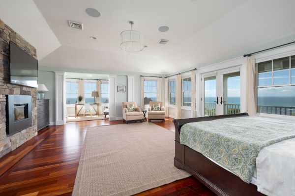 A spacious bedroom with a large bed, wooden floors, fireplace, seating area, and ocean-view windows, creating a serene atmosphere.