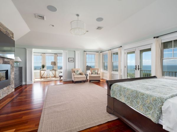 A spacious bedroom with a large bed, wooden floors, fireplace, seating area, and ocean-view windows, creating a serene atmosphere.