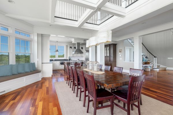 The image shows a spacious, modern dining area with a long table, chairs, and a view into a kitchen, featuring large windows and wood floors.