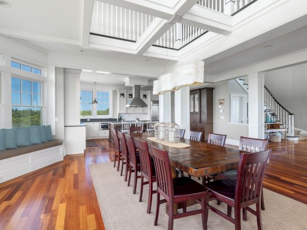The image shows a spacious, modern dining area with a long table, chairs, and a view into a kitchen, featuring large windows and wood floors.