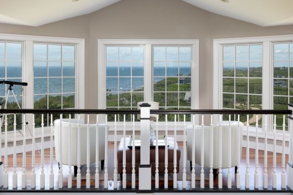A room with large windows offers a panoramic ocean view. Inside are two chairs, a small table, and a telescope.