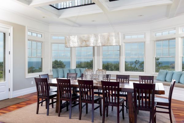 A dining room with a large wooden table, chairs, and a modern chandelier, with large windows offering a scenic view.