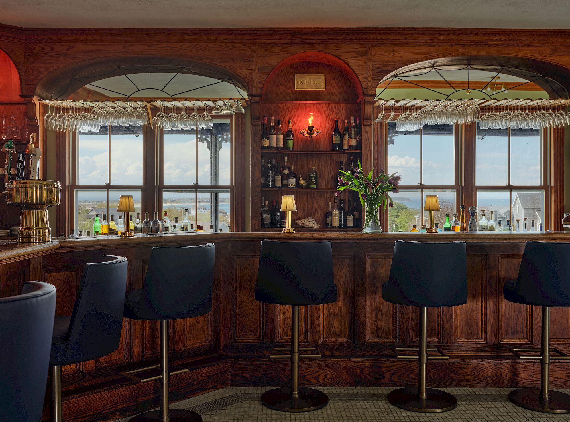 A cozy bar with upholstered stools, stocked shelves, and a view outside through large windows, creating a warm, inviting atmosphere.