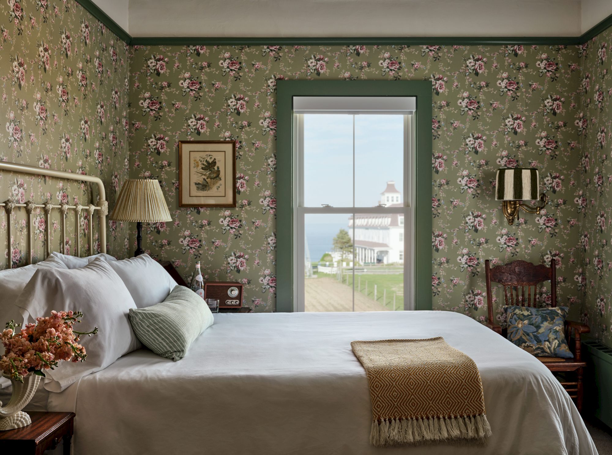 A bedroom with floral wallpaper, a white bed, vintage lamp, and a window view of a scenic building and water outside completes the cozy scene.