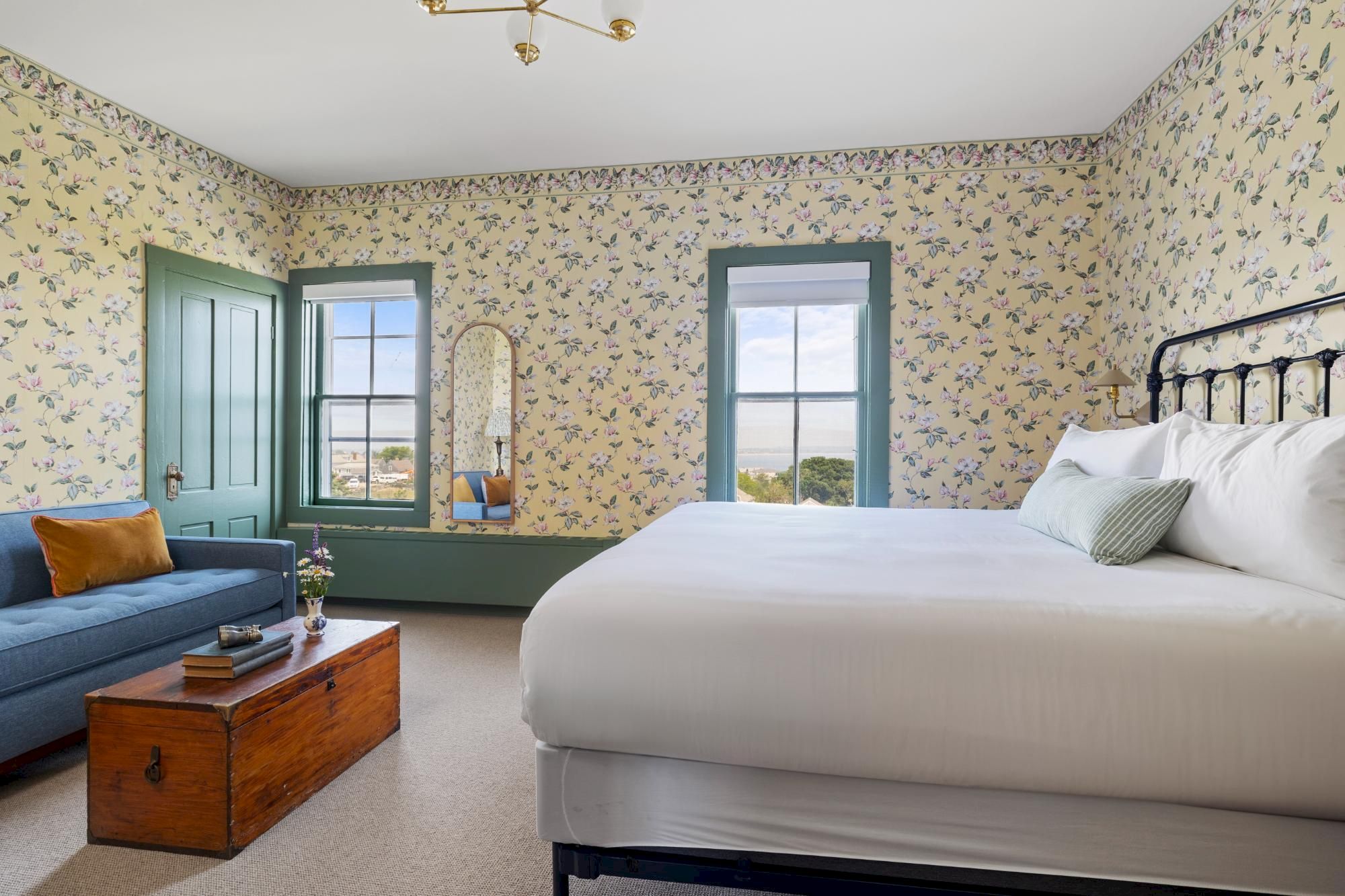 A bright hotel room with a large bed, blue-green trim, floral wallpaper, a blue sofa, a wooden coffee table, and a window view.