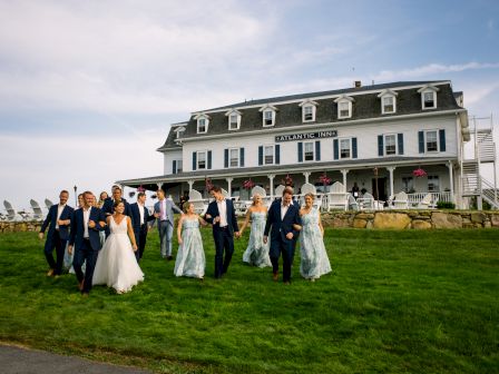 A wedding party walks on a lawn in front of a large, white, historic-looking building with a sign that reads "BLOCK ISLAND INN".