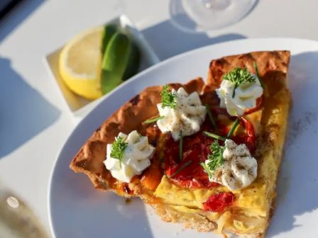 A slice of savory quiche topped with cream and herbs sits on a plate, with lemon and lime wedges in the background.