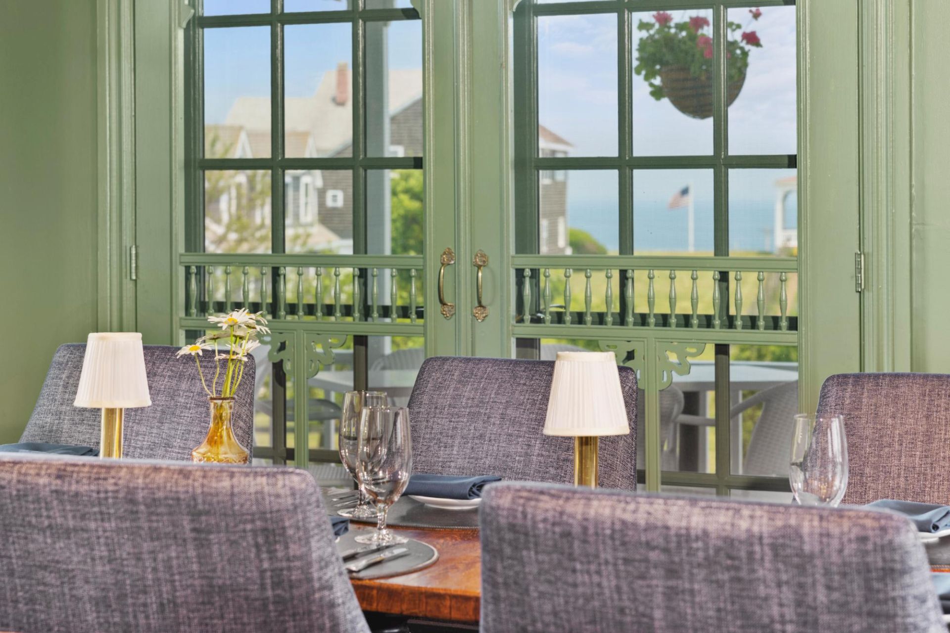 A cozy dining room with a wooden table set for a meal, purple upholstered chairs, table lamps, wine glasses, and a sunny view through large green-framed windows.