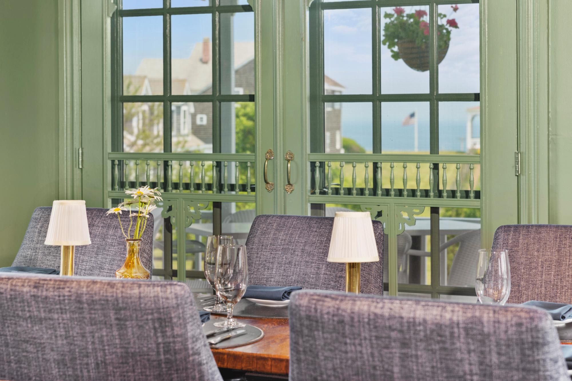 A cozy dining room with a wooden table set for a meal, purple upholstered chairs, table lamps, wine glasses, and a sunny view through large green-framed windows.