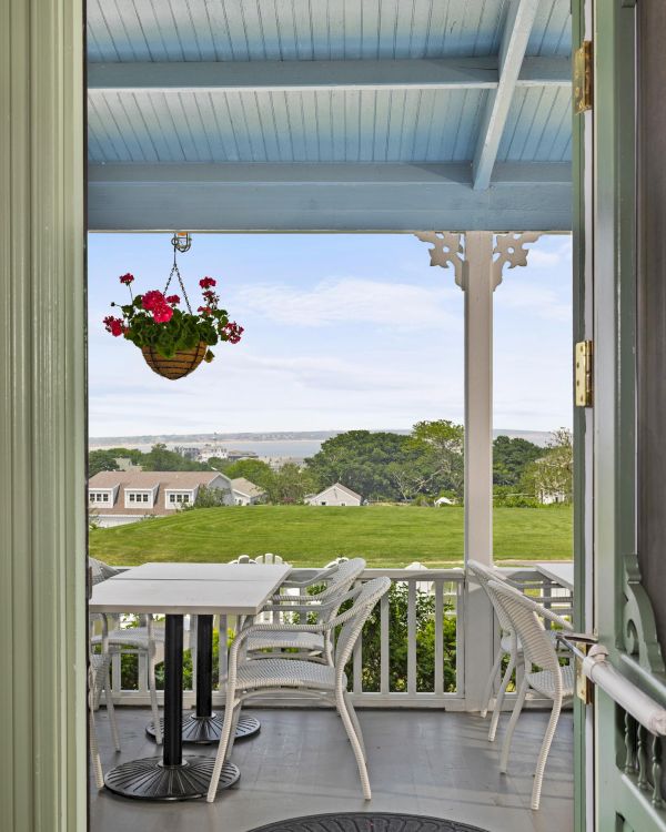 A porch view of a coastal home: hanging basket, white table and chairs, pastel blue ceiling, green garden, and distant sea under a sunny sky.