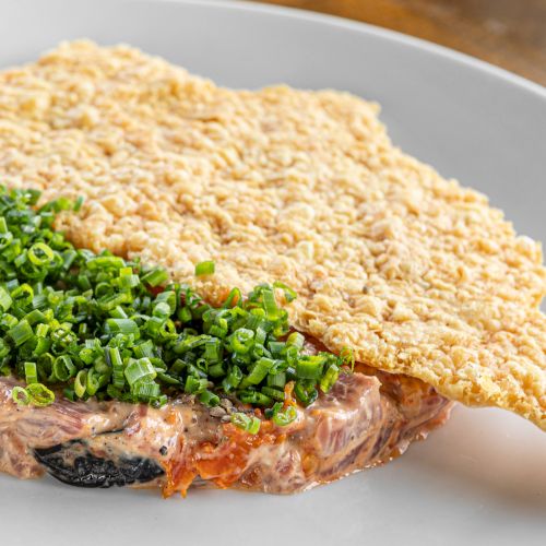 A savory open-faced toast topped with chopped chives, on a slice of meat with a crispy rice cracker perched beside it.
